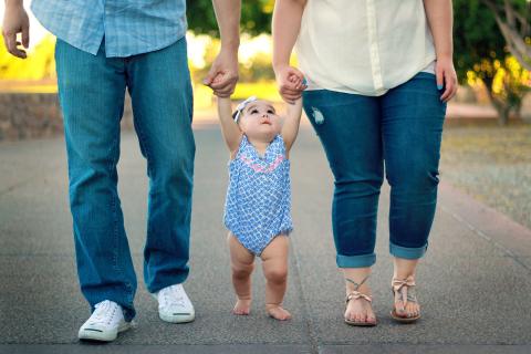 Two parents each hold one of their infant daughter's hands as they help her walk down a path. 