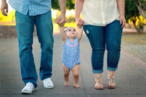 Two parents each hold one of their infant daughter's hands as they help her walk down a path.