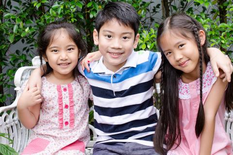 Brother wraps his arms around his two sisters as they sit outside on a bench.