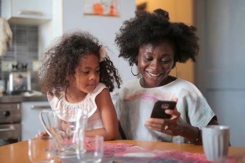 A mother smiles while looking at her cell phone as a young girl looks over her shoulder.