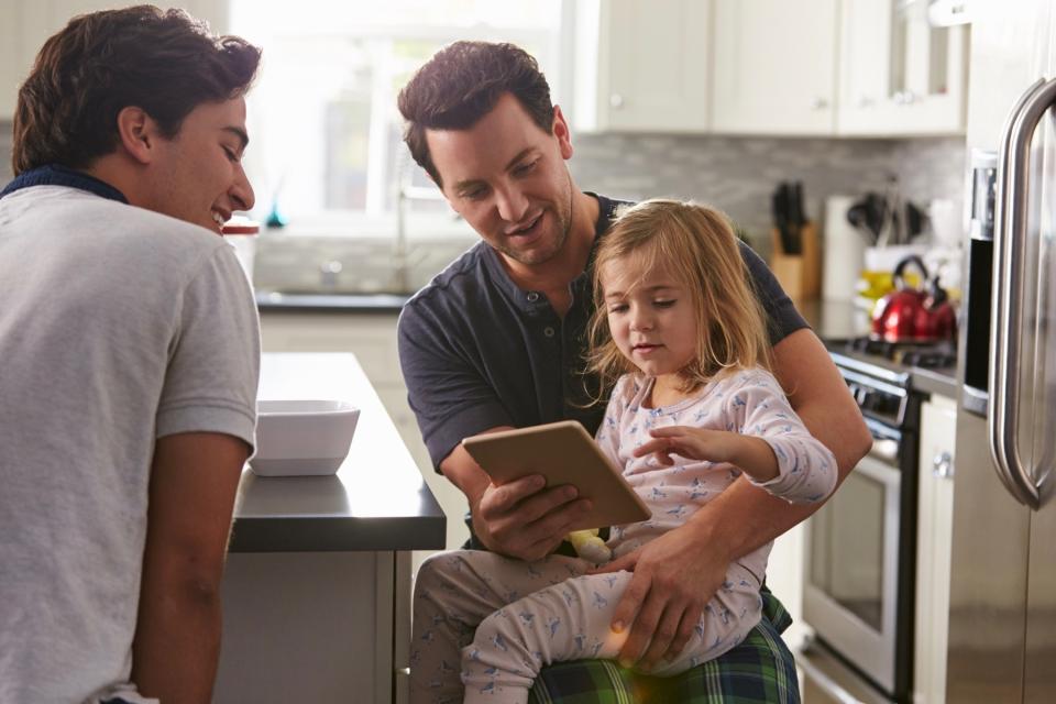 Two dads and their daughter in the kitchen looking at a tablet.