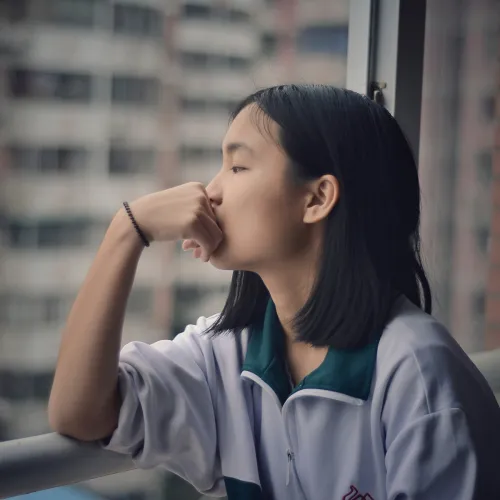 Teenaged girl looks morosely out the window of a high rise apartment