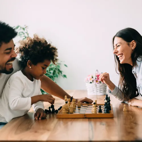 Co-parents play chess with their child together at a dining room table.