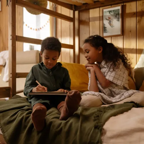 Siblings sit on a bed together while the brother draws.