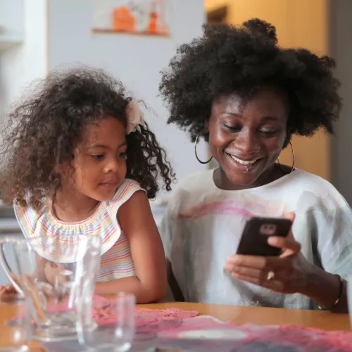 A mother smiles while looking at her cell phone as a young girl looks over her shoulder.