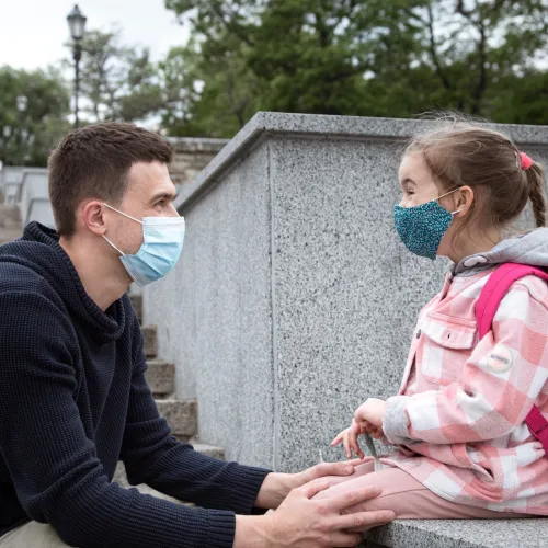 A young girl wearing a face mask and a pink backpack sits on a ledge with her dad hold her knees.