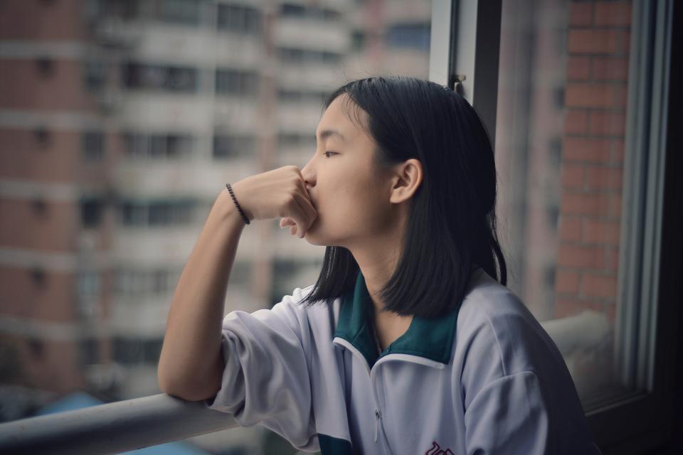 Teenaged girl looks morosely out the window of a high rise apartment