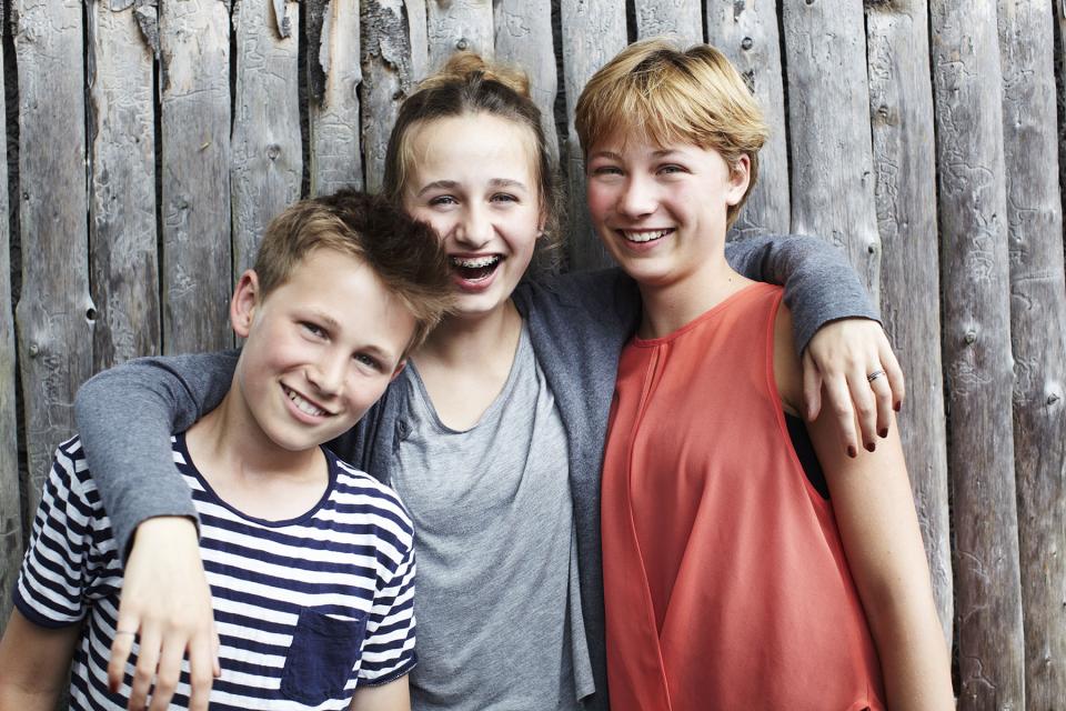 A girl puts her arms around her sister and brother as all three stand laughing as they pose for a picture