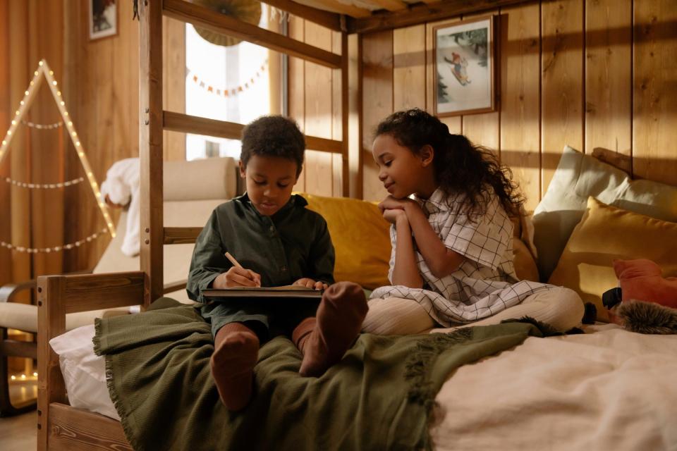 Siblings sit on a bed together while the brother draws.