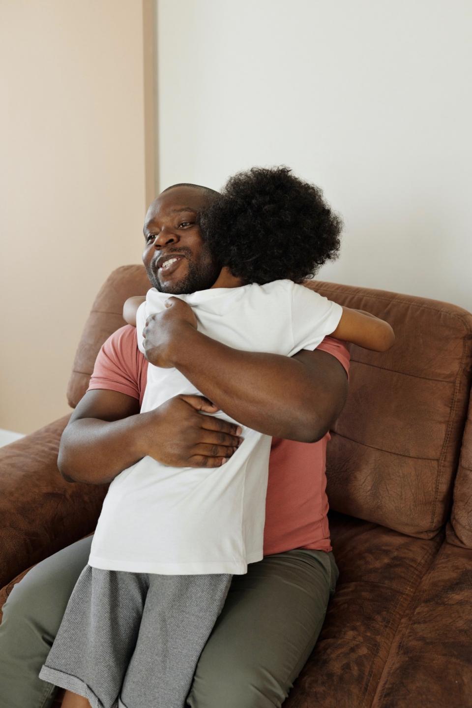 A father sitting in a chair comforts his young child with a hug.