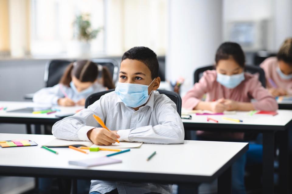 A young boy wears a face mask looks up as he works at a desk in a classroom filled with kids also wearing face masks.