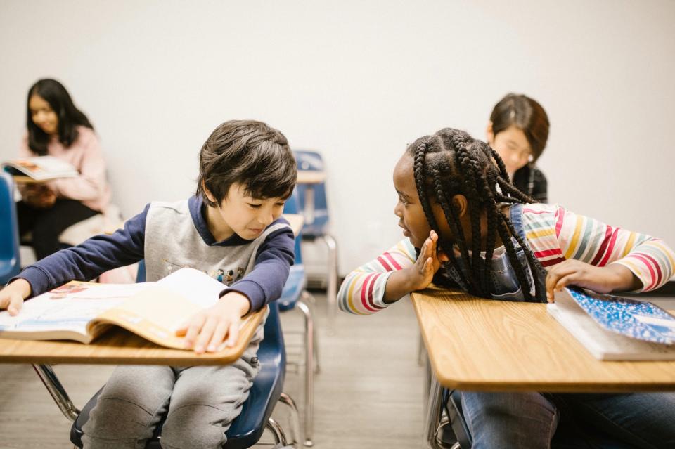 Kids in a classroom. Two sit at desks right next to each other, and they whisper to each other 