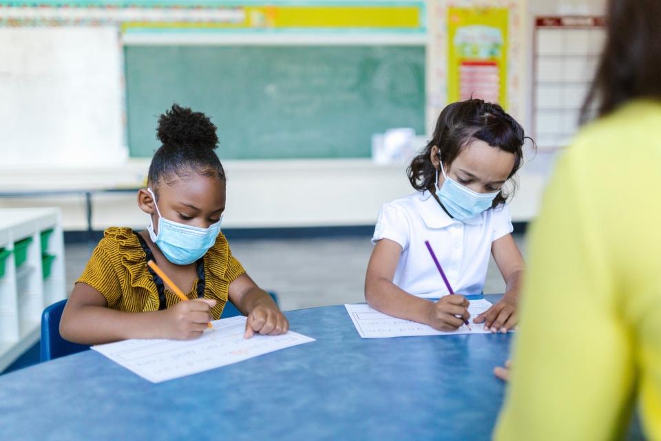 Two young girls wearing face masks sit at a table in a classroom as they both write on separate papers.