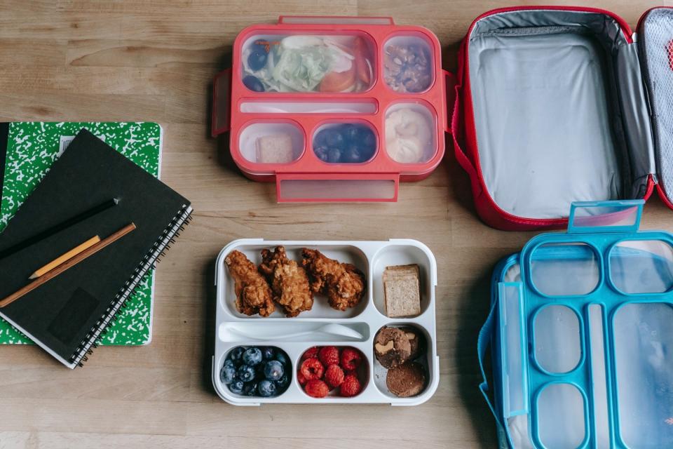 Two lunch boxes and containers filled with food rest on a table next to a stack of books and pencils.