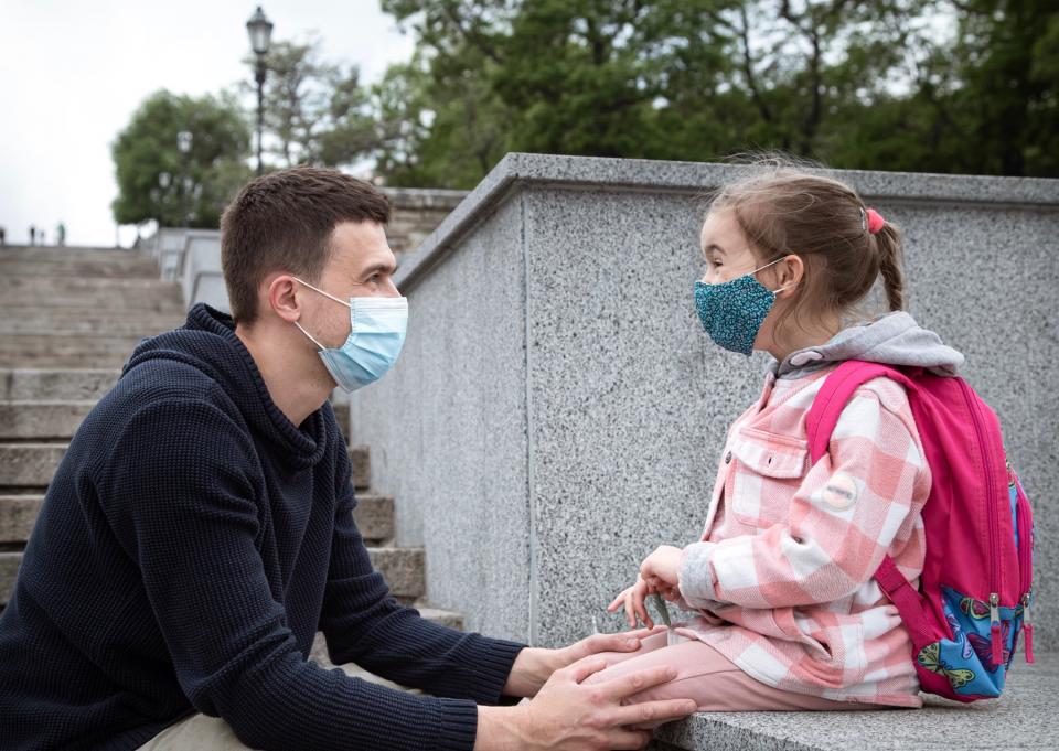 A young girl wearing a face mask and a pink backpack sits on a ledge with her dad hold her knees.