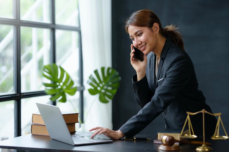 Woman in a suit talks on the phone while working on her computer in front of a window.