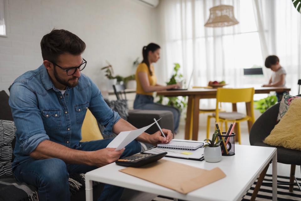 A man sits on a couch to do his taxes while his wife and child sit at a table behind him.