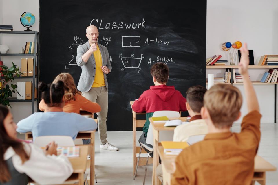 A teacher leading a classroom full of children points to a boy with his hand raised.